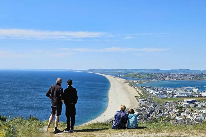 Menschen stehen auf einem grasbewachsenen Hügel und genießen den weiten Blick auf Chesil Beach