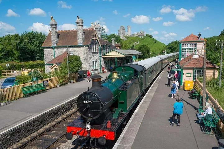Ein historischer grüner Dampfzug, der am Bahnhof von Corfe Castle einfährt, dahinter die Burgruine